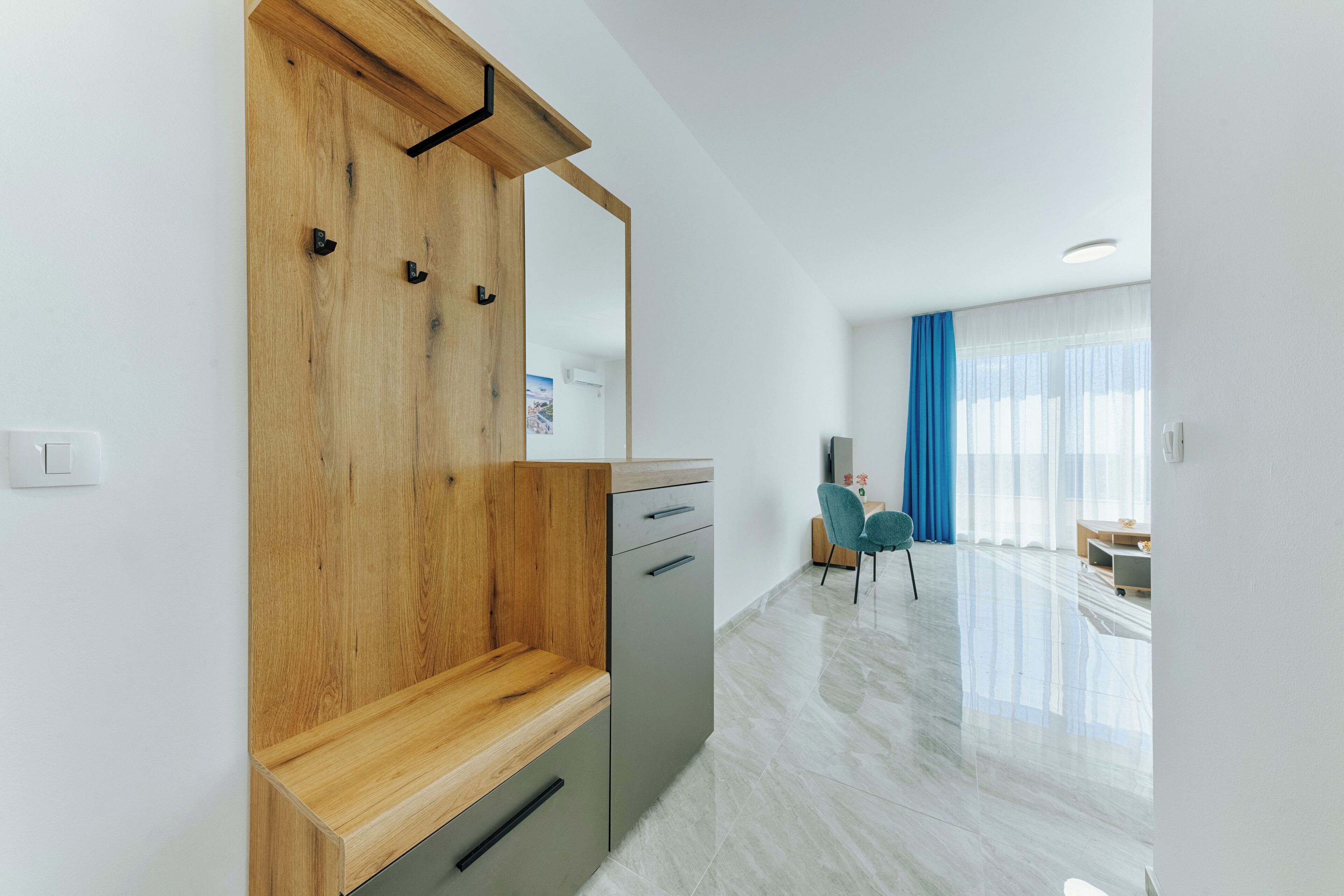 A modern room with a wooden bench, a grey cabinet, a mirror, a blue chair, and a large window with curtains.