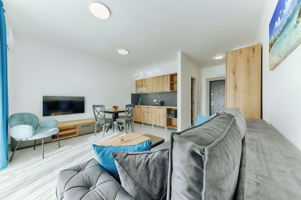 Un salon moderne avec un canapé gris, des coussins bleus et gris, un téléviseur à écran plat, un coin repas avec des chaises, une cuisine avec des meubles en bois et un tableau accroché au mur.