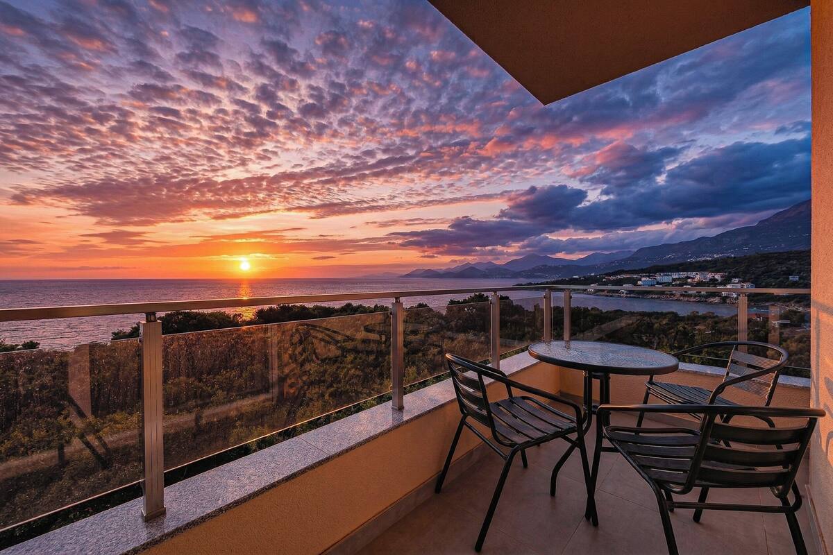 Un balcon avec une table et des chaises, d’où l’on peut admirer un coucher de soleil sur la mer et les collines lointaines.