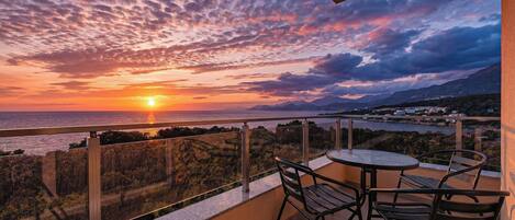 A balcony with a table and chairs, overlooking a sunset over the sea and distant hills.