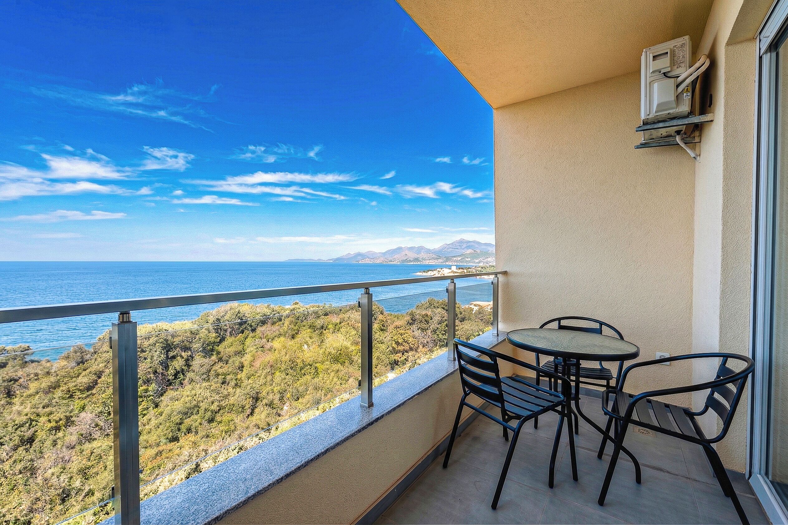 Un balcon avec une table et des chaises, donnant sur un paysage côtier avec des arbres et des montagnes.