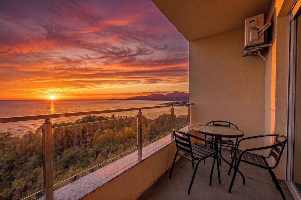 Un balcon avec une table et des chaises, d’où l’on peut admirer un coucher de soleil sur une étendue d’eau et des montagnes.