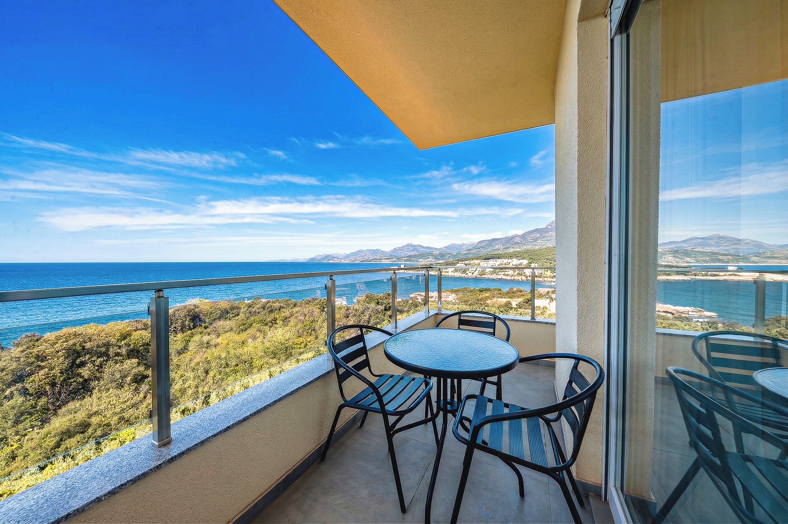 A balcony with a table and chairs, overlooking a coastal landscape with mountains and a body of water.