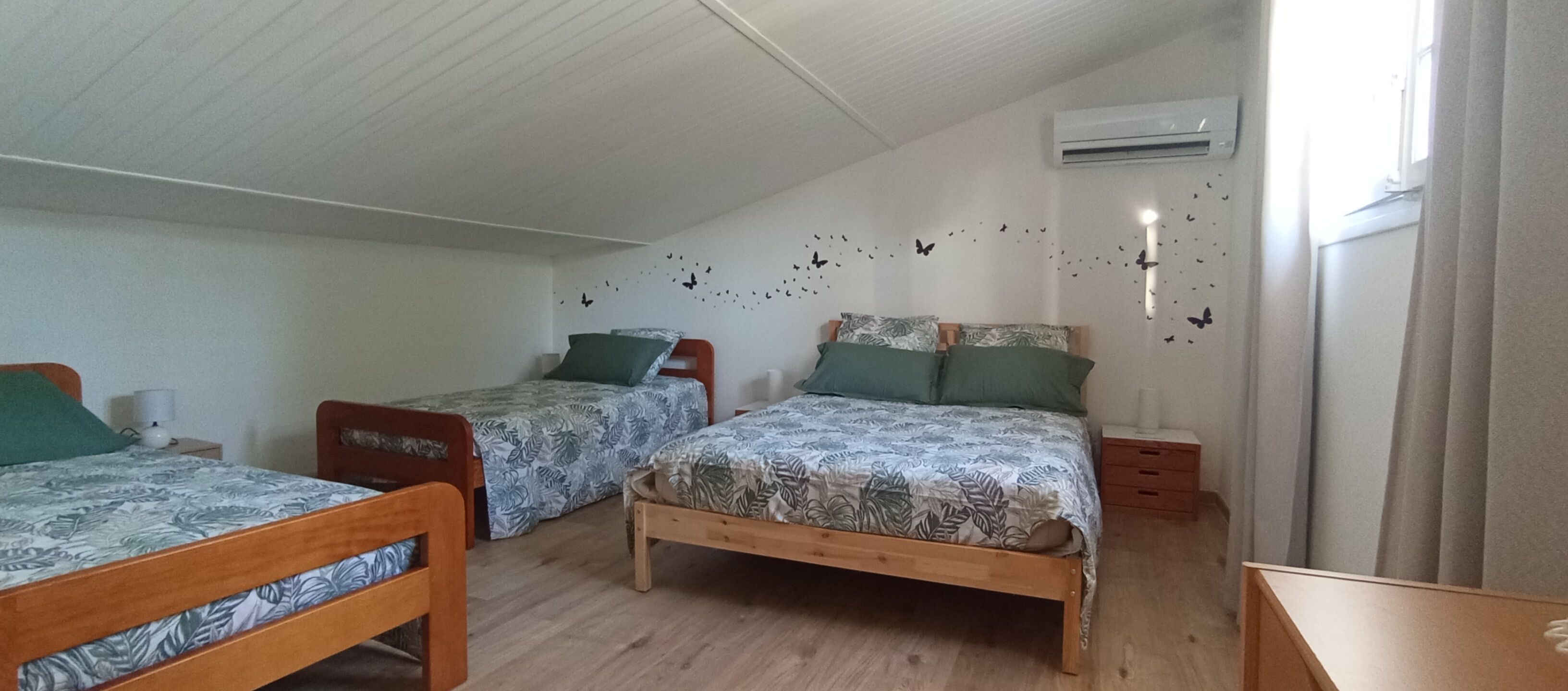 A room with two single beds, a wooden floor, a wall-mounted air conditioner, and a window with curtains.