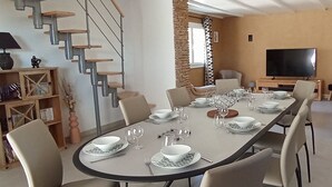 Ein Essbereich mit einem langen Tisch, gedeckt für sechs Personen, umgeben von Stühlen, einer Couch und einem Fernseher im Hintergrund.