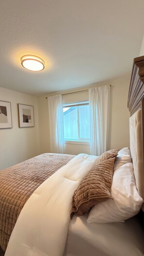A neatly made bed with a quilted headboard, a window with curtains, and framed artwork on the wall.