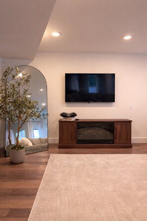 A living room with a flat-screen TV mounted on the wall, a wooden entertainment center, a large mirror, and a potted plant.