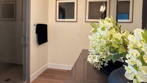 Un baño moderno con cabina de ducha de vidrio, tocador con espejo y un florero con flores blancas.