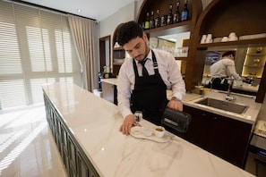 Un barista preparando café en una barra de mármol en una habitación luminosa, con un ventanal y vistas a la ciudad.