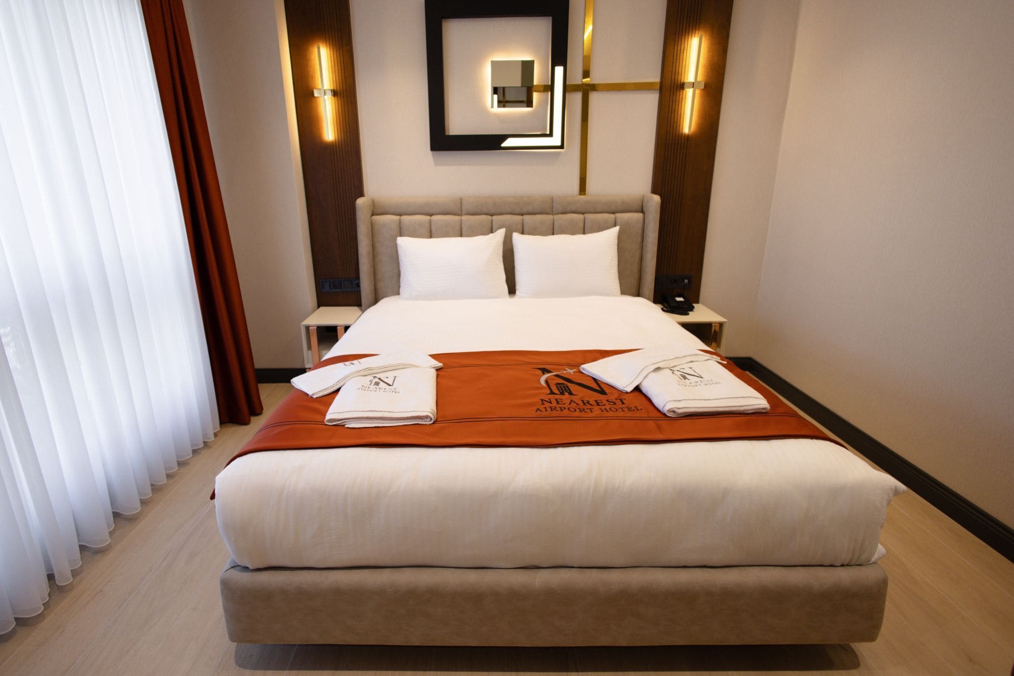 A hotel room with a large bed, a nightstand, a wall-mounted light, and a window with curtains.