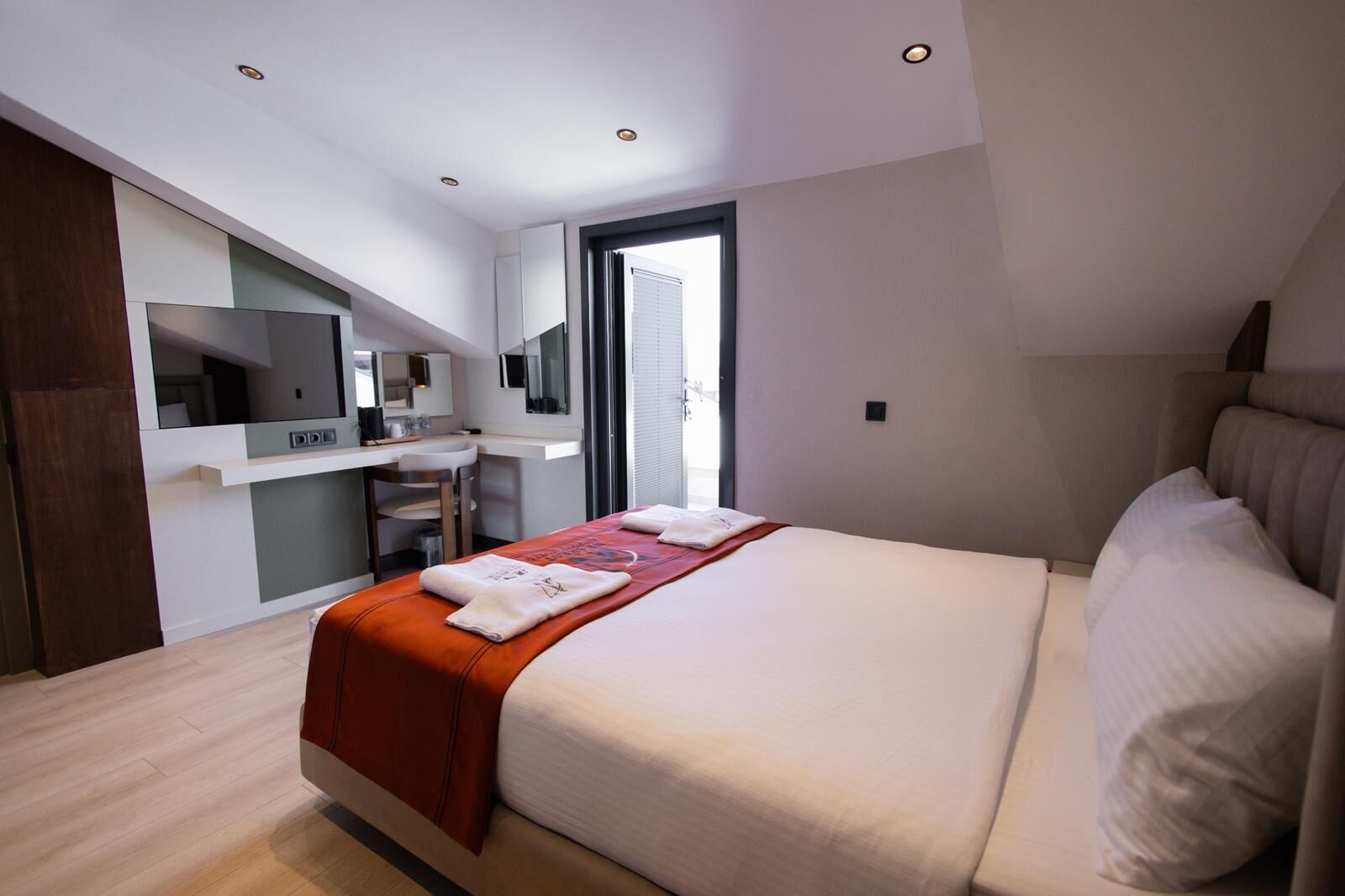 A modern hotel room with a large bed, a desk with a chair, a mirror, and a window.
