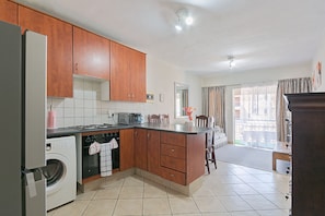 A kitchen with wooden cabinets, a washing machine, and a dining area with a table and chairs. - Cozy Midrand Stay with Pool , Wi-Fi , Secure Parking&Self Checkin. (Midrand)