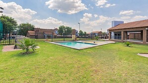 Una piscina rodeada de césped, una valla, un edificio de ladrillo y un cielo despejado.