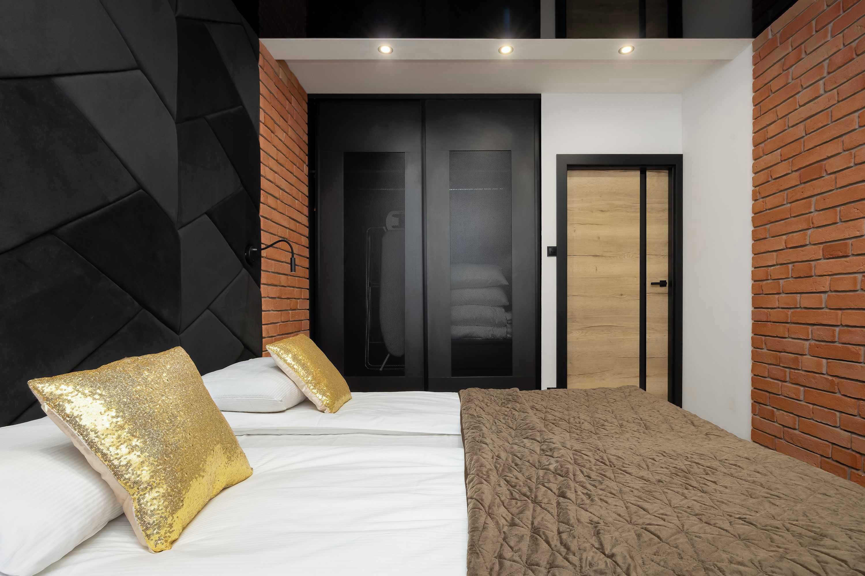 A modern bedroom with a bed, pillows, a quilted blanket, a black headboard, a wooden door, and brick walls.