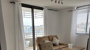 A modern living room with a sofa, armchair, glass coffee table, and a view of the city through large windows.