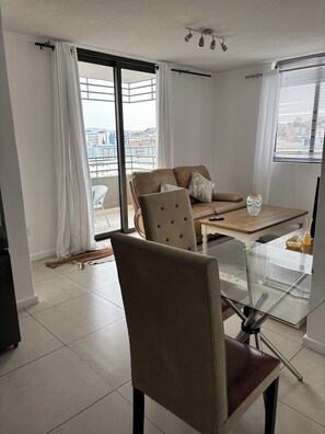A modern living room with a sofa, armchair, glass coffee table, and a view of the city through large windows. - Cozy CBD Apartment - Windhoek (Windhoek)