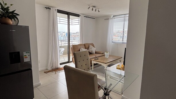 A modern living room with a glass dining table, a sofa, a chair, and a refrigerator.