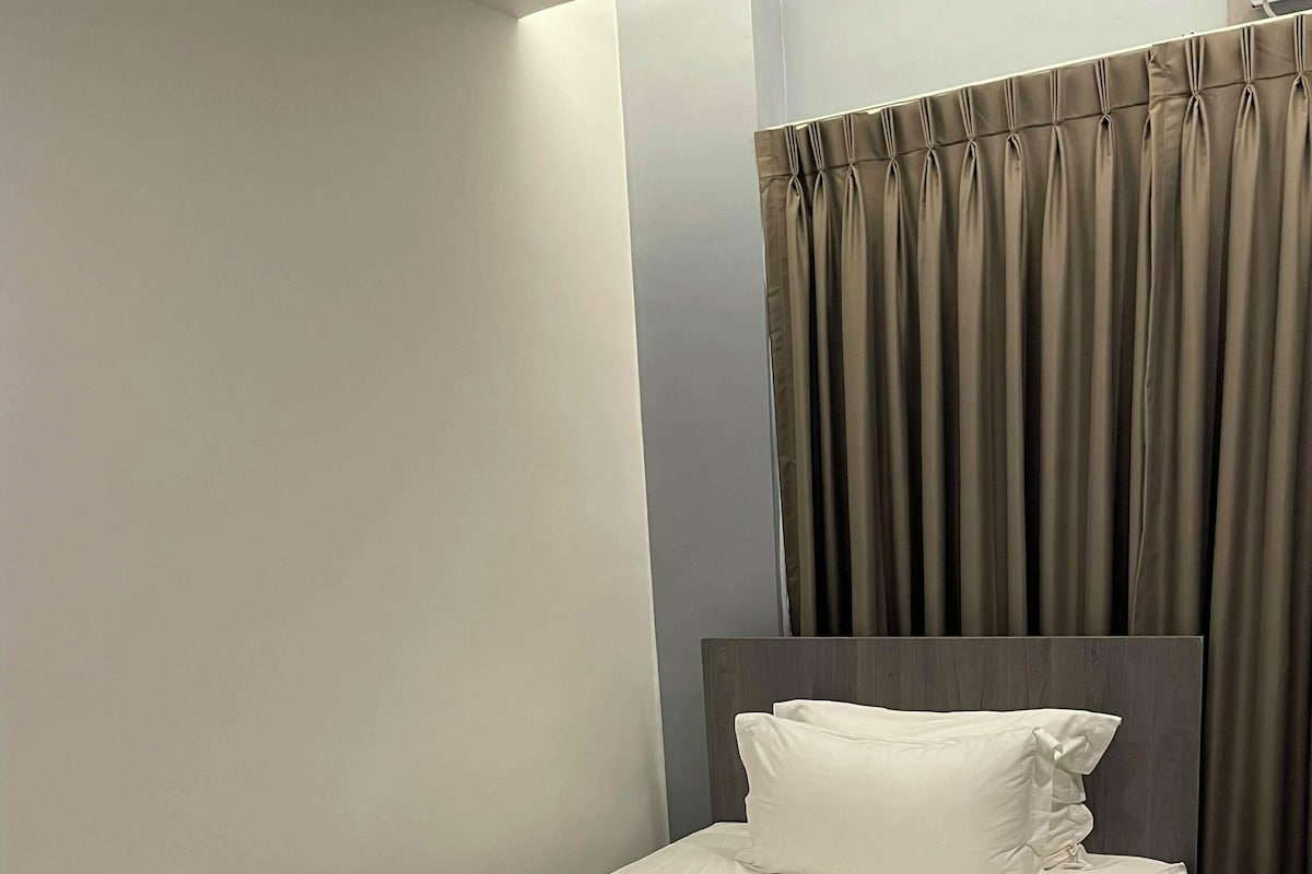 A neatly made bed with white linens and a folded towel, a dark headboard, and a window with curtains.