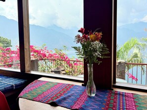 A table with a colorful woven tablecloth, a clear vase with flowers, and a view of mountains and pink flowers through large windows. - Tiny Cabin #4 Lake Atitlan (San Juan la laguna,)