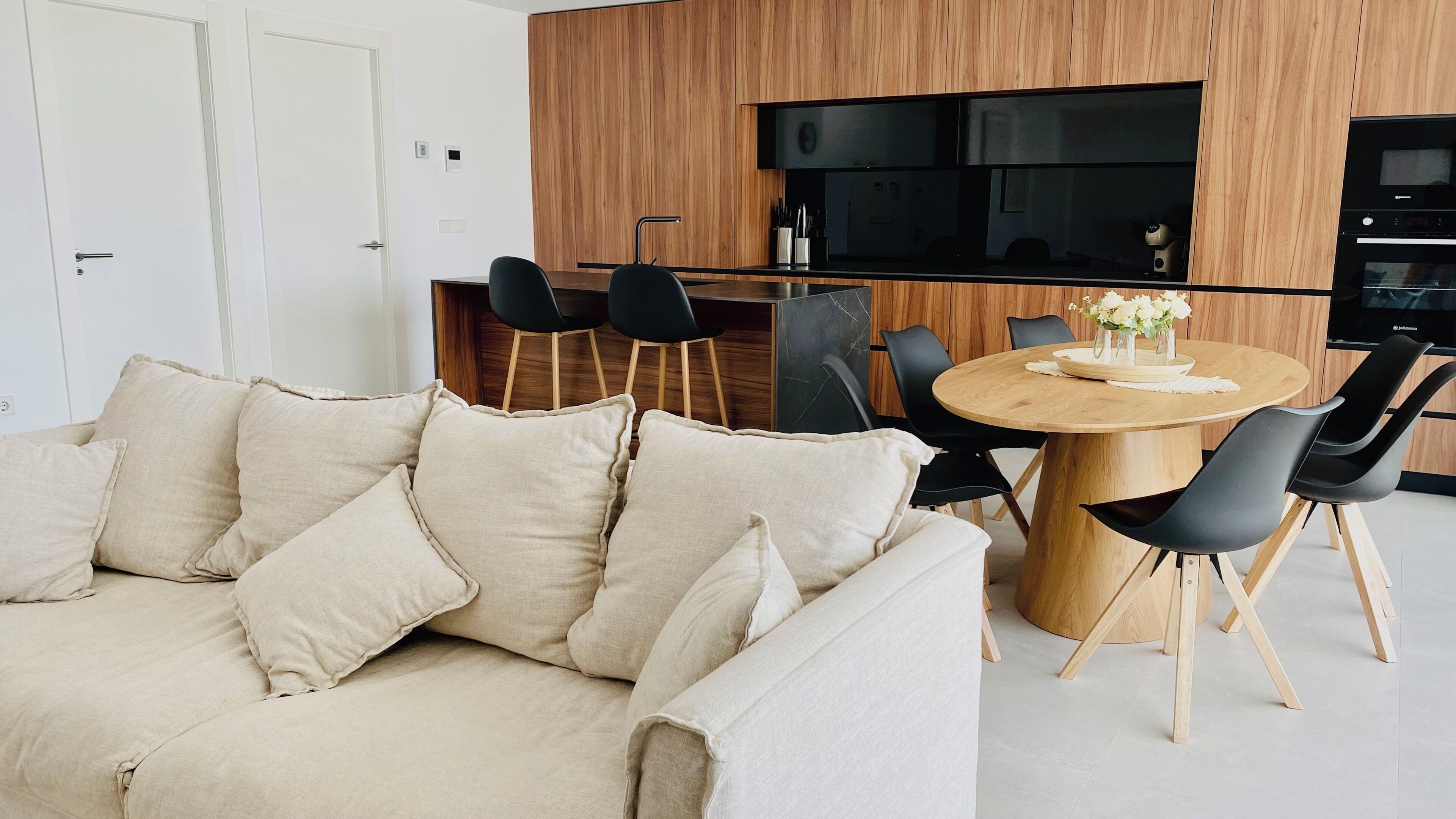 A modern living room with a beige sofa, a wooden dining table with black chairs, and a kitchen area with wooden cabinets and a black countertop.