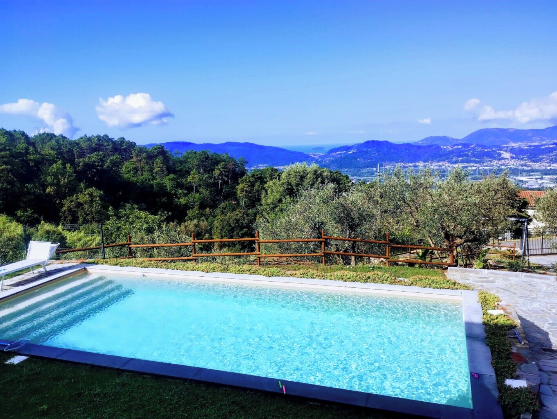 Ein rechteckiger Swimmingpool mit Holzzaun, umgeben von Gras und Bäumen, mit Blick auf Berge und eine Stadt in der Ferne.