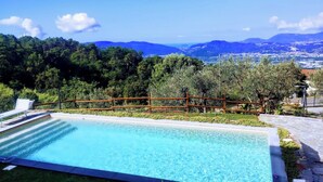 A rectangular swimming pool with a wooden fence, surrounded by grass and trees, with a view of mountains and a town in the distance. - Villa Terrace Vert, 12 seats, Saltwater pool, A/C, gym, BBQ (Sarzana)