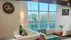 A reception area with a curved reception desk, a clock, a sofa, and a large window with a view of trees and a building. - HOTEL NATRAJ (Sirsa)