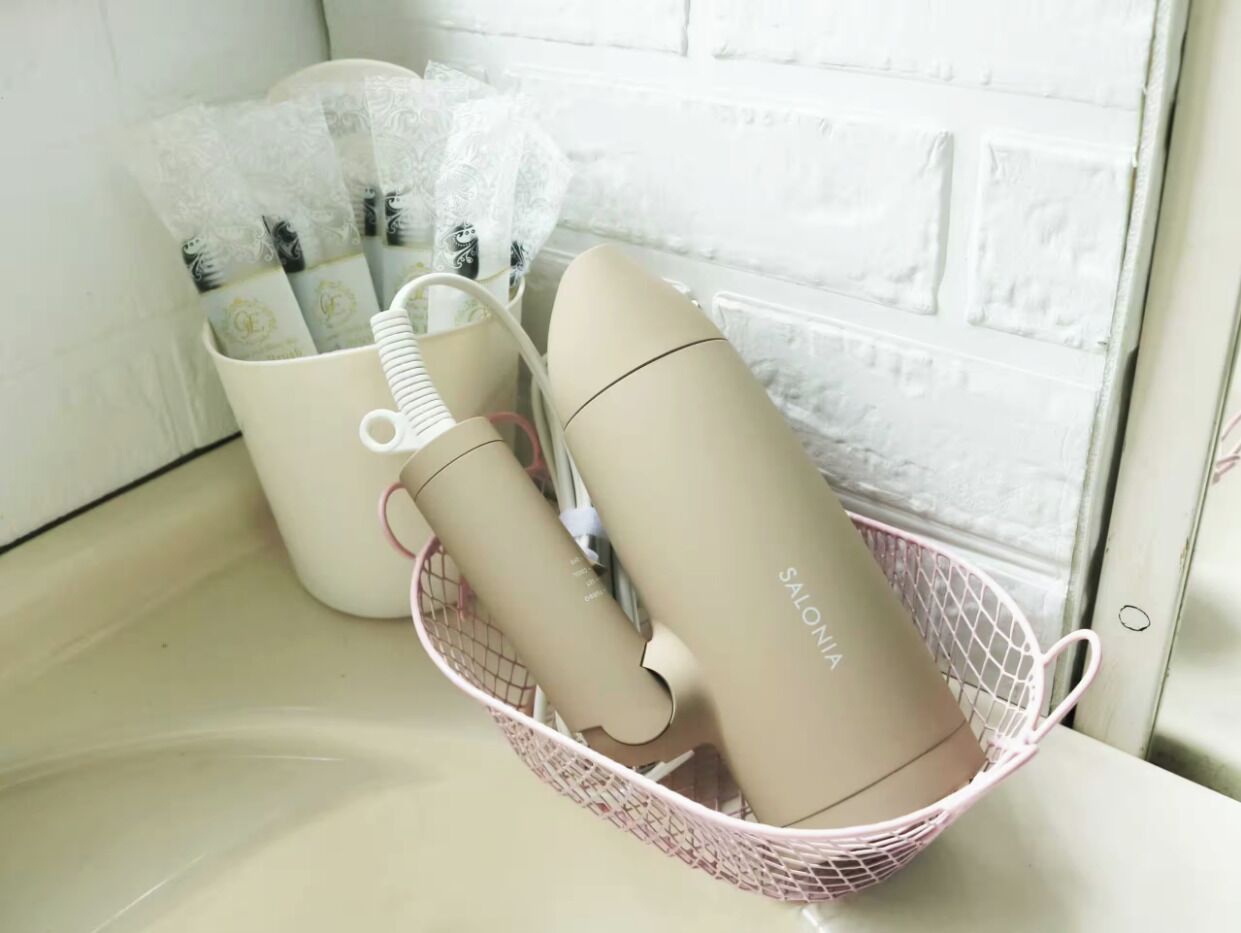 Un muro de ladrillo blanco, un contenedor color beige con la palabra 'SALONIA' escrita y una cesta de alambre rosa que contiene un objeto blanco.