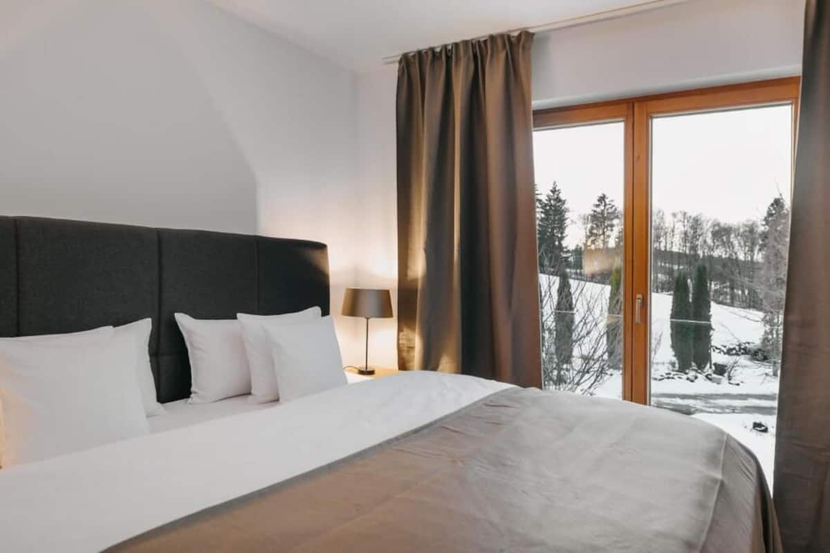 Ein Hotelzimmer mit einem großen Bett, einem Nachttisch mit Lampe und Blick auf eine verschneite Landschaft durch eine Schiebetür.
