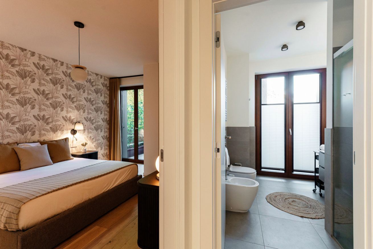 A bedroom with a bed, bedside table, and a nightstand. There is a bathroom with a toilet, bidet, and a shower area.