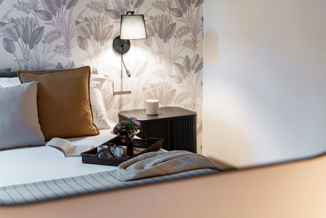 A neatly made bed with pillows, a bedside table with a lamp, a tray with a cup and a book, and a floral wallpaper.