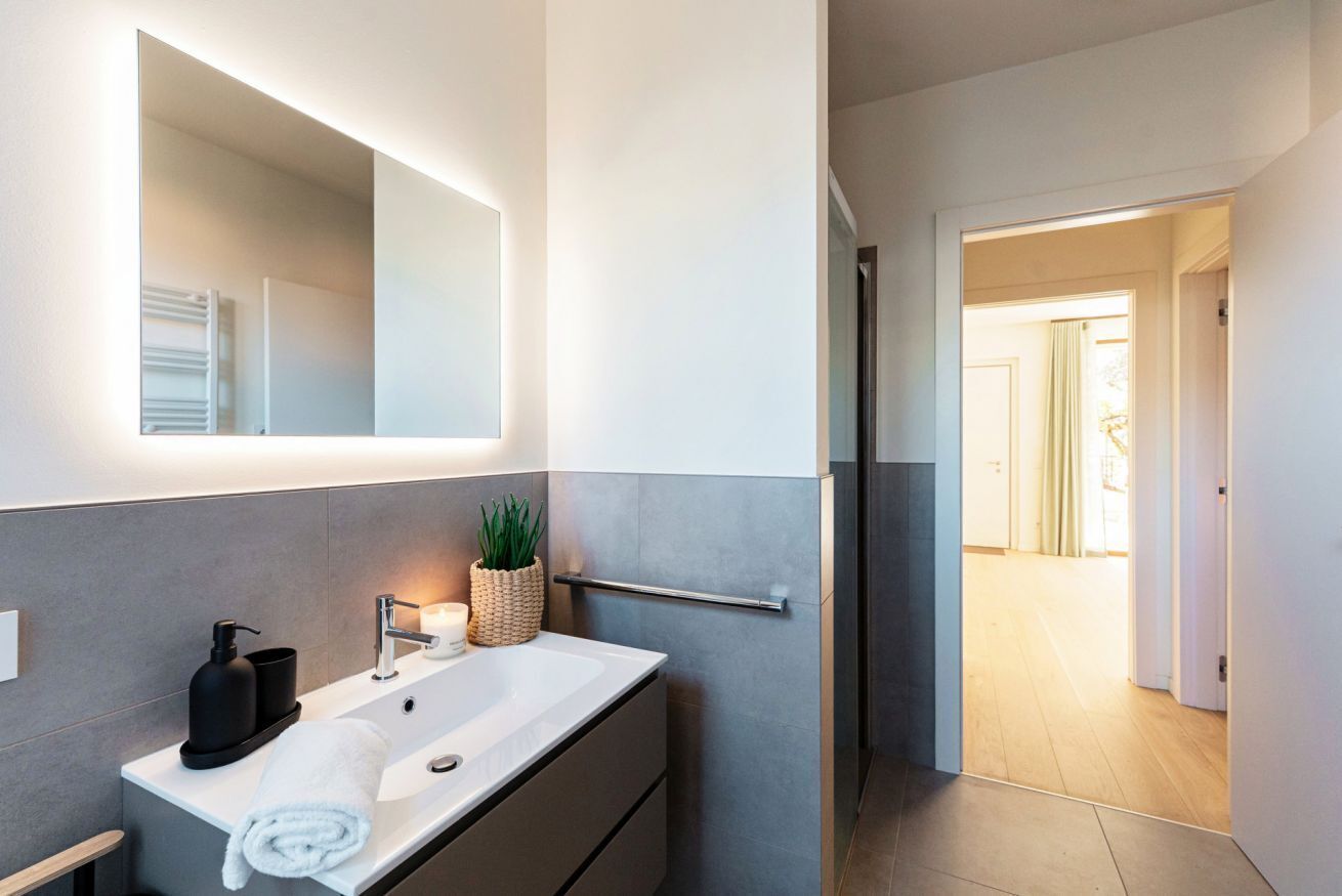 Een moderne badkamer met een grote spiegel, een witte wastafel, een handdoek, een zeepdispenser en een potplant.