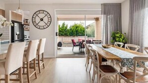 A modern kitchen with a clock, a dining area with a table and chairs, and a sliding glass door leading to an outdoor patio. - Luxury Sunbury retreat (Sunbury)