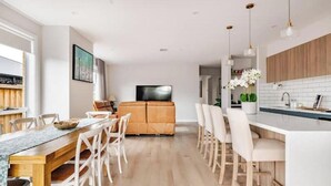 A modern kitchen with a central island, wooden cabinets, and a dining area with white chairs. - Luxury Sunbury retreat (Sunbury)