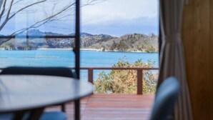 A room with a view of a lake and hills, a round table, and a wooden deck.
