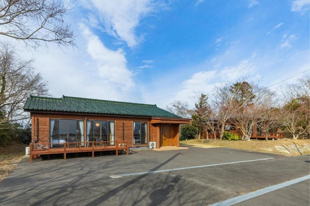 A wooden cabin with a green roof, large windows, and a covered outdoor seating area.