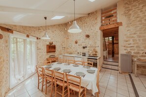 A dining area with a stone wall, wooden furniture, and a kitchen in the background. - Gite authentique de charme Rocamadour (Trespoux-Rassiels)