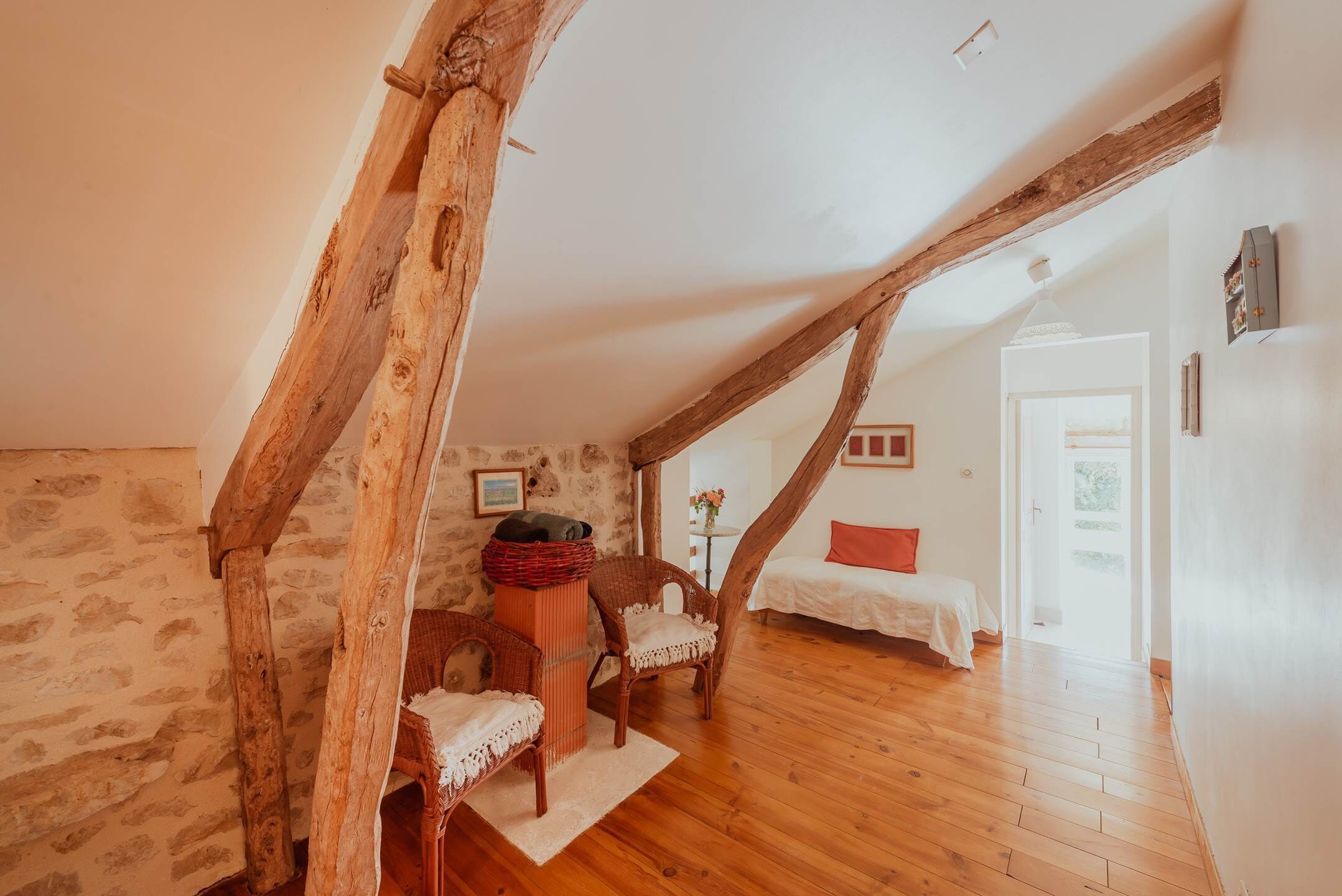 A room with exposed wooden beams, a bed with a red pillow, a wicker chair, and a small table with a basket.
