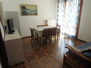 A dining area with a table and chairs, a television on a stand, a painting on the wall, and patterned flooring. - Apartment 'Residence Torino 29' with Shared Pool and Private Terrace (Lido di Jesolo)