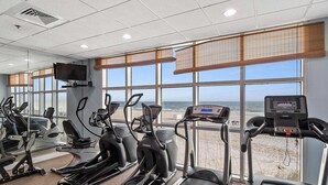 Ginásio com esteiras, bicicletas ergométricas e uma tela de televisão, com vista para a praia.