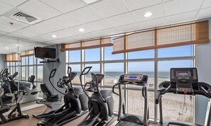 A gym with treadmills, stationary bikes, and a TV screen, overlooking a beach. - Gulf-Front Corner 3BR • Wrap Balcony • Sleeps 10 (Gulf Shores)