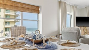Quarto com vista para o oceano, televisão de tela plana, móveis de vime e uma mesa posta com jogos americanos e objetos decorativos.