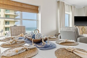 A room with a view of the ocean, a flat-screen TV, wicker furniture, and a table set with placemats and decorative items. - Gulf-Front Corner 3BR • Wrap Balcony • Sleeps 10 (Gulf Shores)