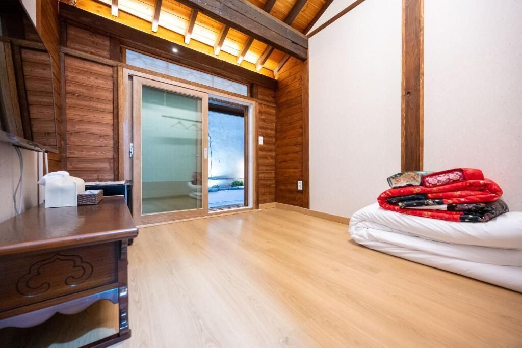 Habitación con paredes de madera, puerta corredera de vidrio, cómoda de madera y cama con colcha roja y negra.