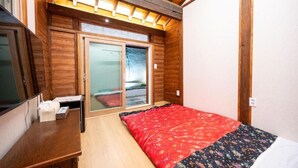Ein Zimmer mit Holzwänden, einem Bett mit rot-schwarz gemustertem Überwurf, einem Holztisch und einer Schiebeglastür, die zu einem Außenbereich führt.