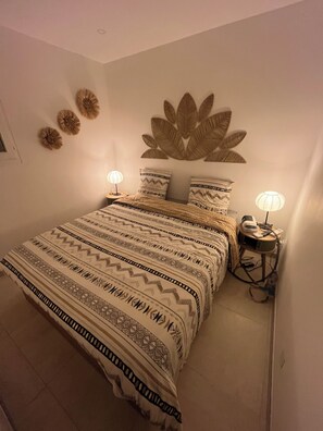 Ein ordentlich bezogenes Bett mit gemusterten Betttextilien, zwei Nachttischlampen, eine an der Wand befestigte dekorative Blattform und ein kleiner runder Tisch mit einer Lampe.