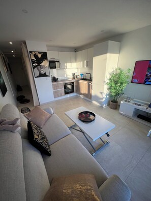 Ein modernes Wohnzimmer mit einer Couch, einem Couchtisch und einem Fernseher. Im Hintergrund ist eine Küche mit Schränken und Geräten zu sehen.
