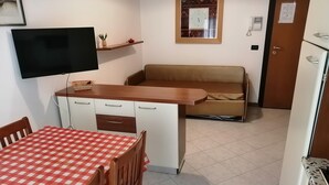 A kitchenette with a TV, a dining table with a checkered tablecloth, a sofa, and a small shelf with decorative items. - Apartment 'Acapulco Residence 201' with Shared Pool and Private Terrace (Lido di Jesolo)