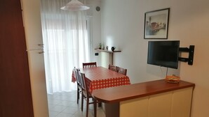 A dining area with a table covered in a checkered tablecloth, wooden chairs, a television mounted on the wall, and a window with sheer curtains. - Apartment 'Acapulco Residence 201' with Shared Pool and Private Terrace (Lido di Jesolo)