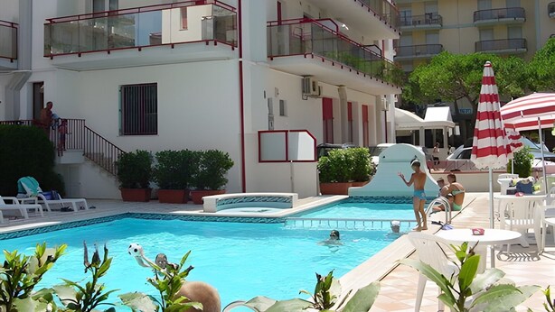 A multi-story hotel with a swimming pool, outdoor seating, and a red and white striped umbrella. - Apartment 'Acapulco Residence 201' with Shared Pool and Private Terrace (Lido di Jesolo)